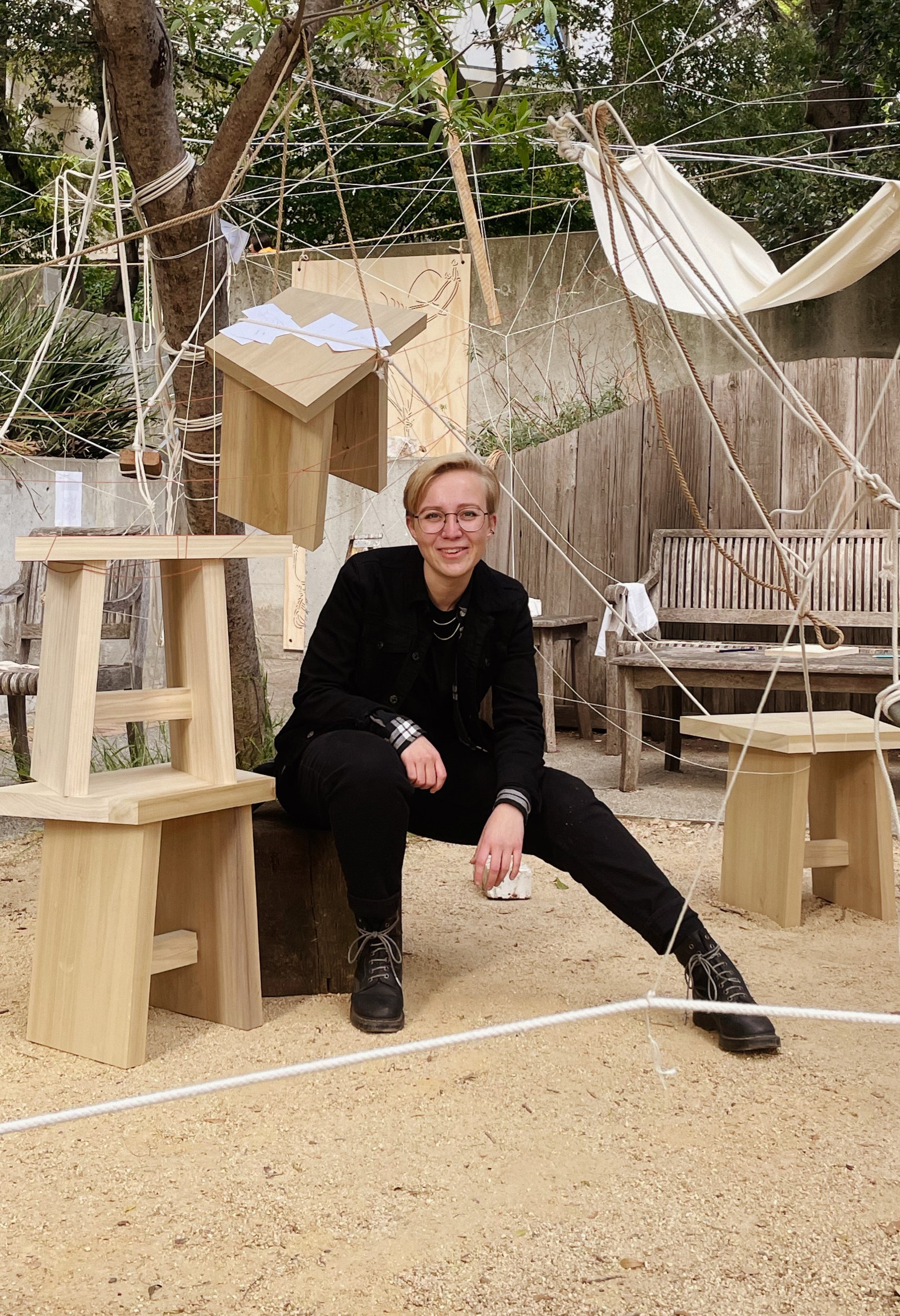 A photograph of Ari smiling at the camera while in a courtyard strung up with textiles, stools, rope, and thread created by the attendees of their thesis exhibit earlier that afternoon